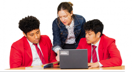 Two students in red blazers are seated with a book and computer on table, A teacher is leaning over between them.